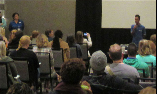 Picture shows a teenaged student standing with a microphone and speaking to a large audience.  A woman sitting in front is videotaping him with her smartphone.